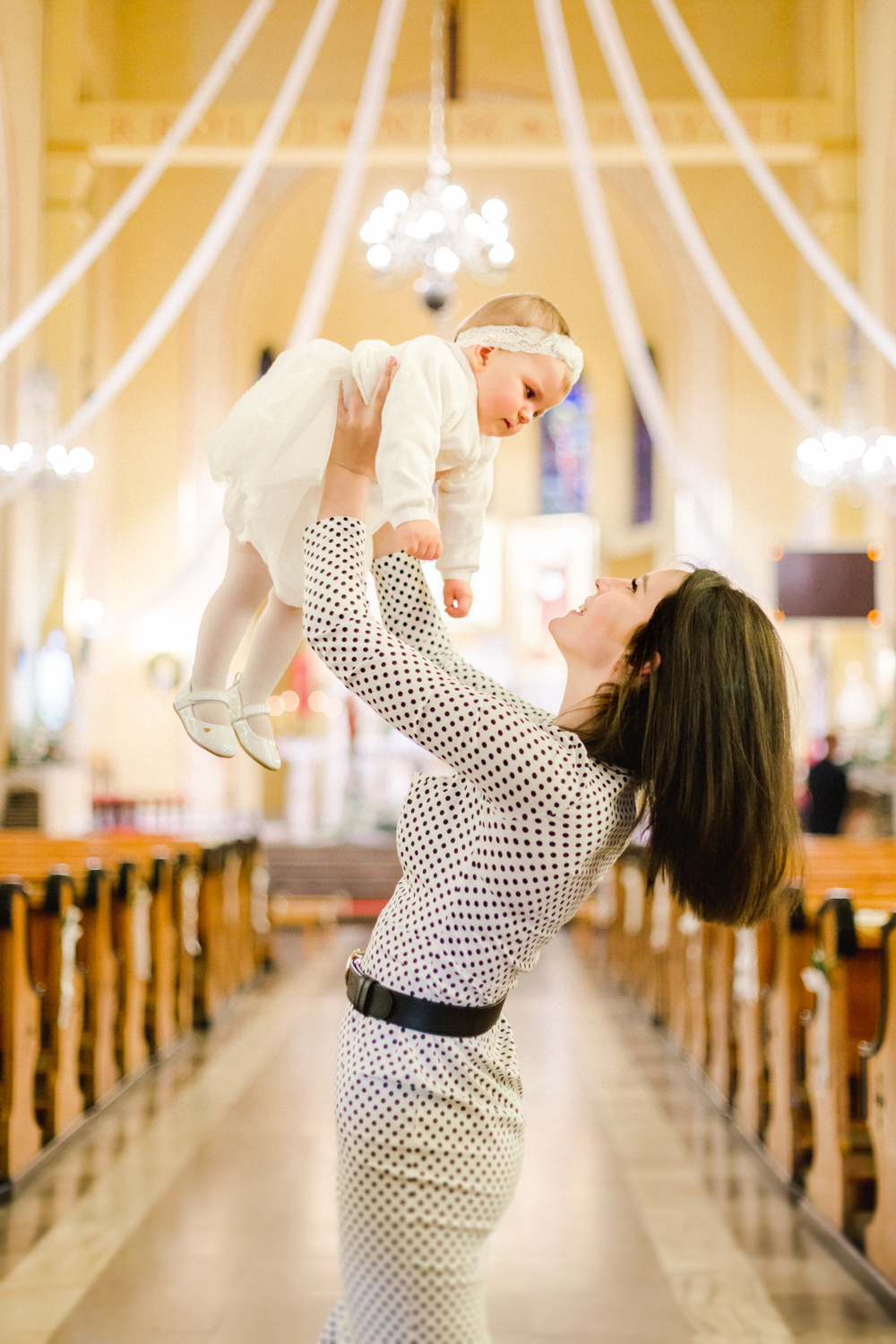 Fotografia mamy podnoszącej niemowlę w eleganckiej sukience podczas chrztu w kościele.