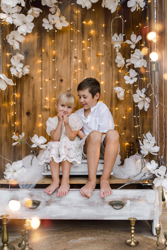 Dwójka uśmiechniętych dzieci pozuje na tle drewnianej ścianki ozdobionej białymi kwiatami i światełkami, idealnej na dziecięcą sesję zdjęciową w Żywcu.