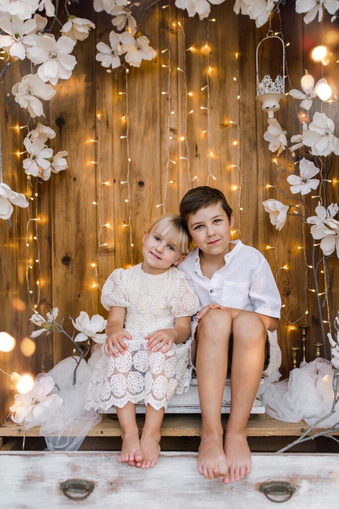 Dziecięca sesja zdjęciowa Żywiec - rodzeństwo, dziewczynka i chłopiec, siedzący na tle drewnianej ścianki z białymi kwiatami i światełkami, uśmiechający się.