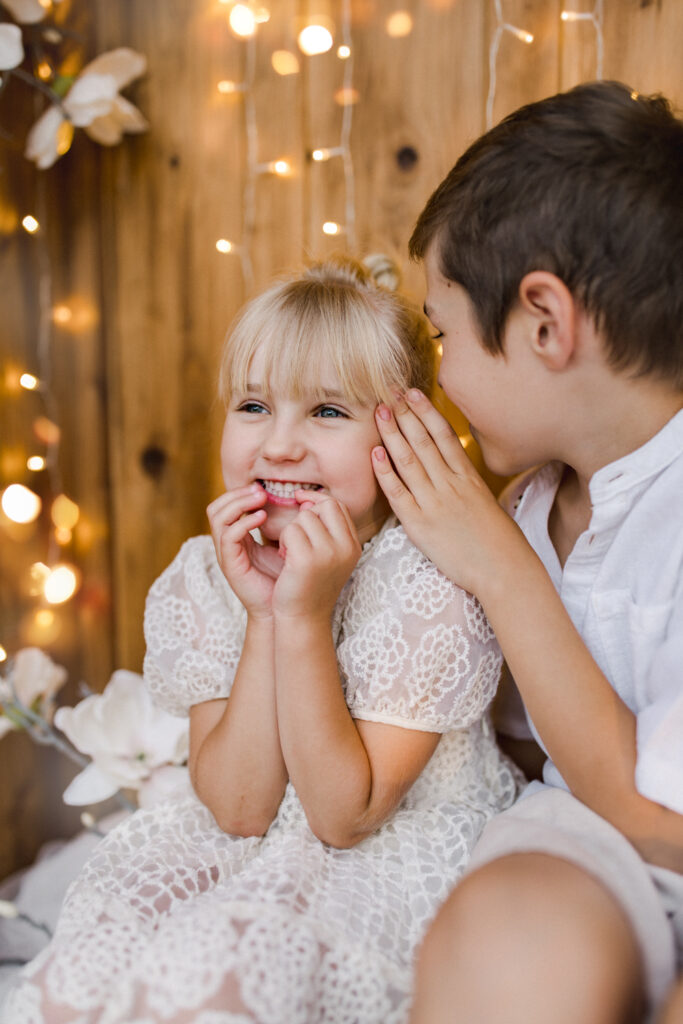 Dziecięca sesja zdjęciowa Żywiec: uśmiechnięte rodzeństwo na tle drewnianej ścianki ze światełkami i kwiatami.